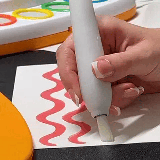Eine Hand mit gepflegten Nägeln zeichnet mit einem weißen Stift rote, wellenförmige Linien auf ein Blatt Papier; im Hintergrund sind bunte Ringe und Malutensilien zu sehen.