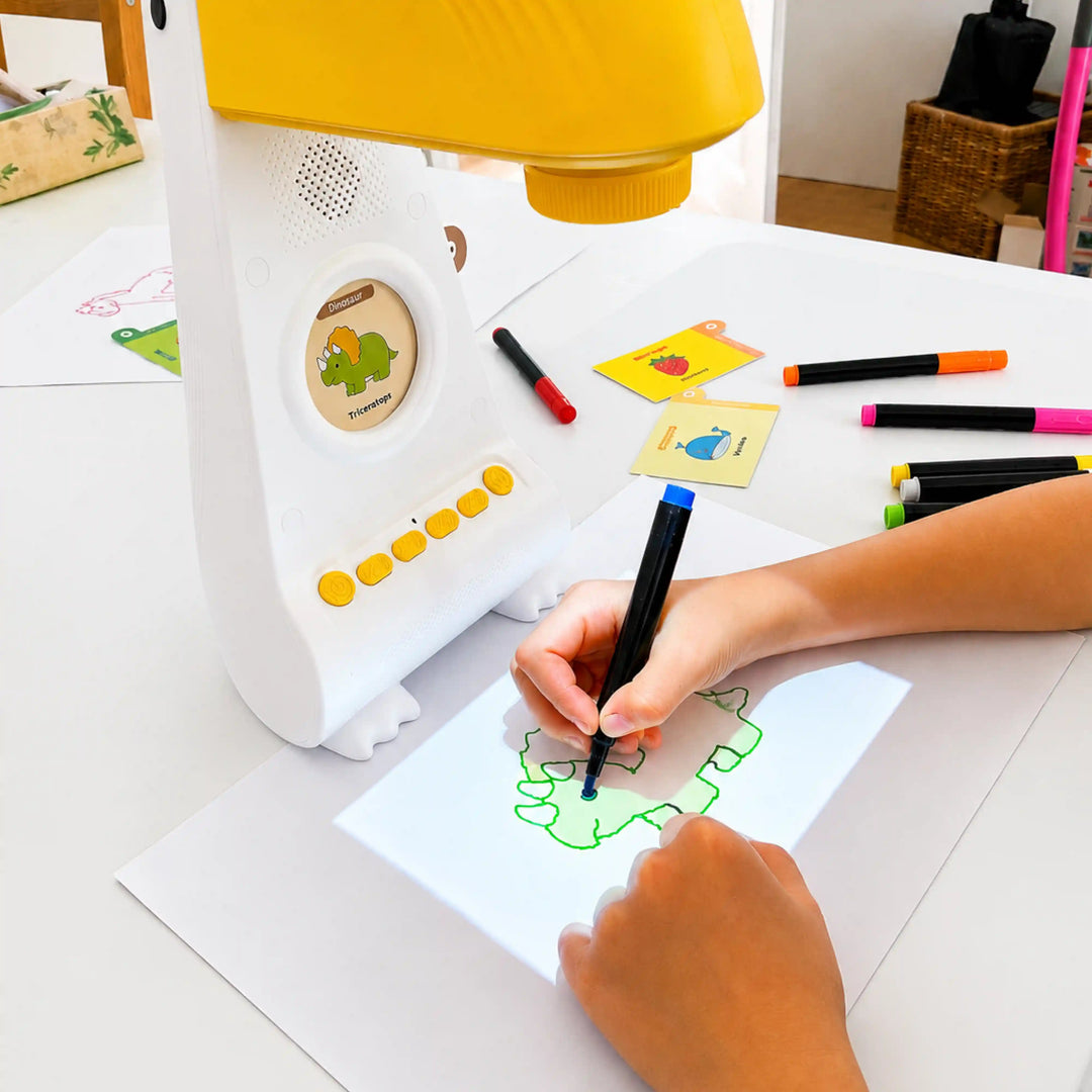 Ein Kind zeichnet mit einem schwarzen Filzstift den Umriss eines grünen Comic-Dinosauriers nach, der von einem gelb-weißen Zeichenapparat auf Papier projiziert wird. Auf dem weißen Tisch liegen bunte Filzstifte und Zeichenkarten.